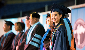 Smiling female graduate in cap and gown at ceremony with faculty in academic robes behind her.