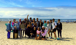 Group of diverse young people posing and smiling on a sunny beach with ocean and sky in background.