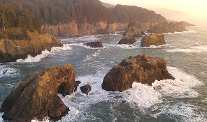 Rocky coastline with waves crashing against large boulders at sunset with forested cliffs in background.
