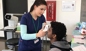 Nurse using a handheld device to check a seated young man's eye in a clinical setting.